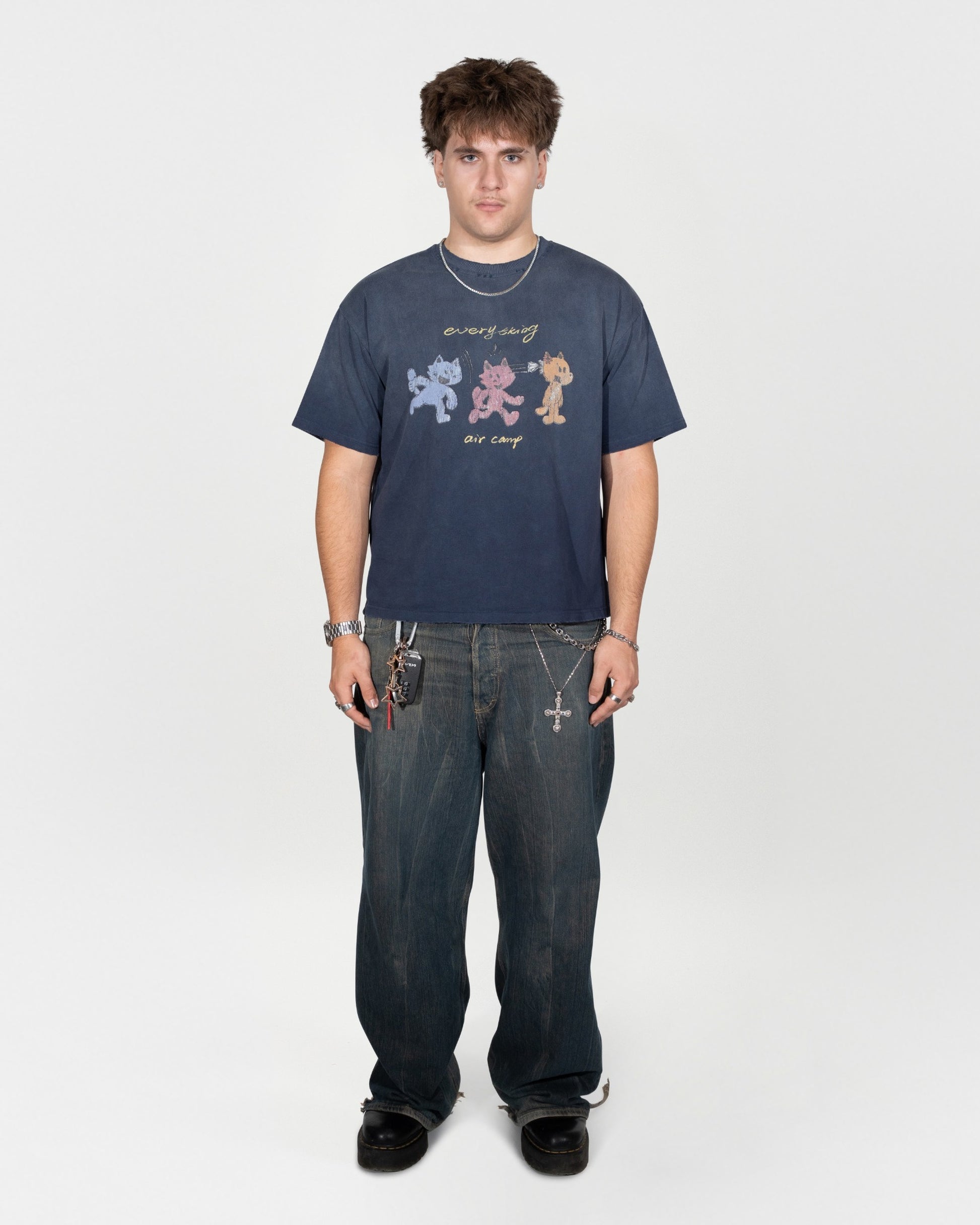 Full body 45 degree angle shot. The model is facing directly at the camera. serious facial expression with hands down by his hips. He is wearing the air camp tee by j3t with dark greyish blue jeans and black shoes.
