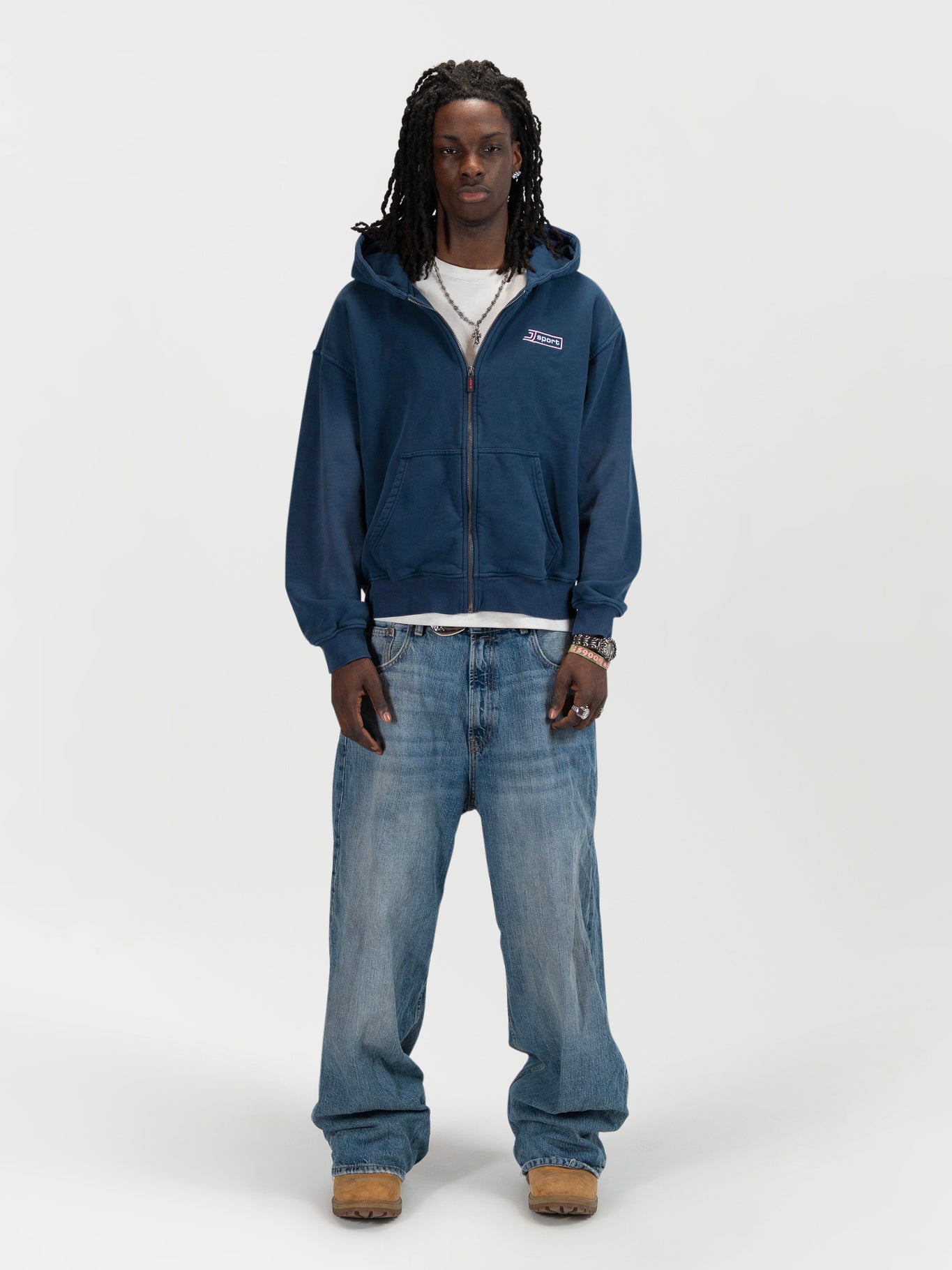 Front facing full body shot of a male model wearing the J Sport zip by J3T. HE has on a white shirt underneath, light blue jeans and light brown boots to complete the fit.