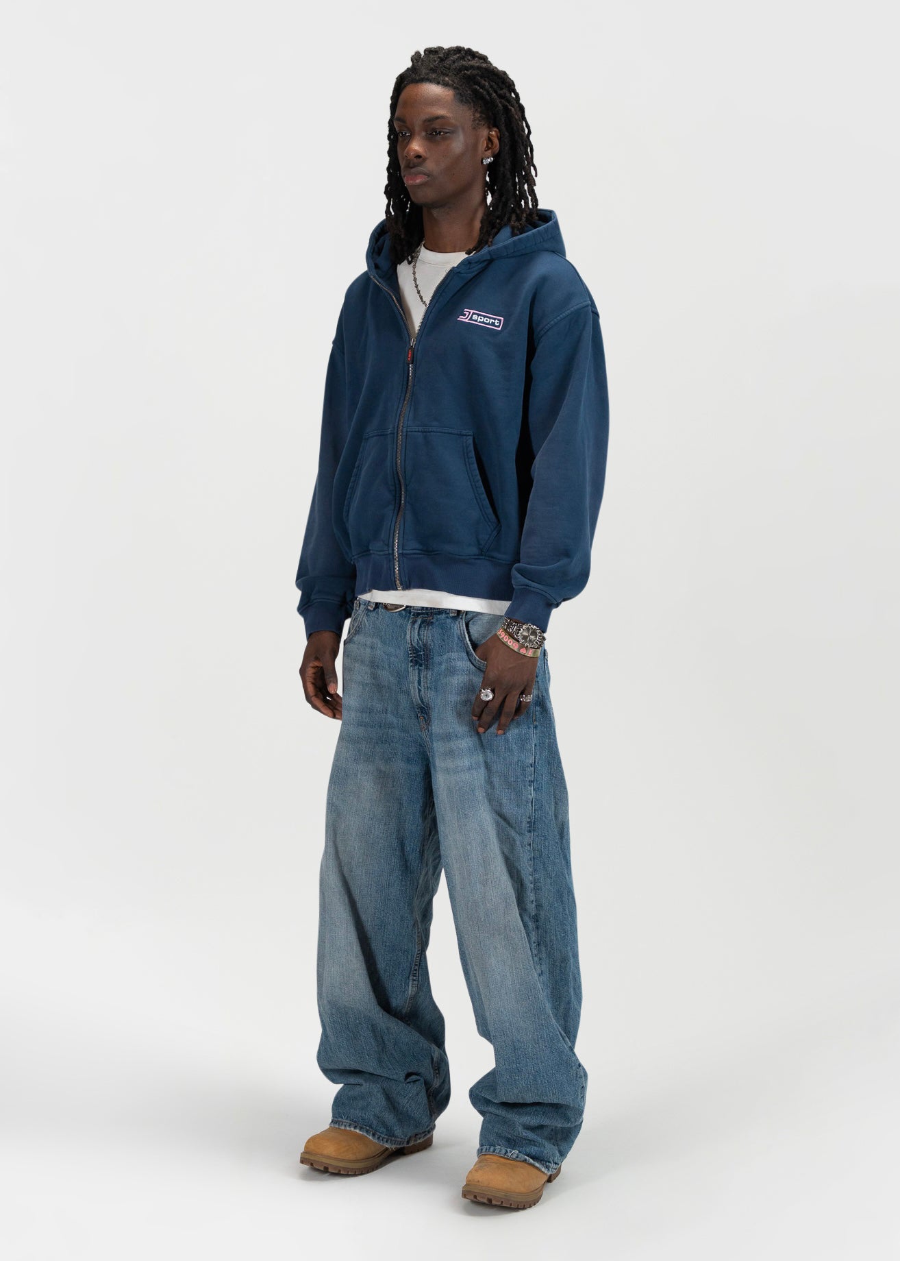 Left 45 degrees front facing full body shot of a male model wearing the J Sport zip by J3T. HE has on a white shirt underneath, light blue jeans and light brown boots to complete the fit. everythingj3t
