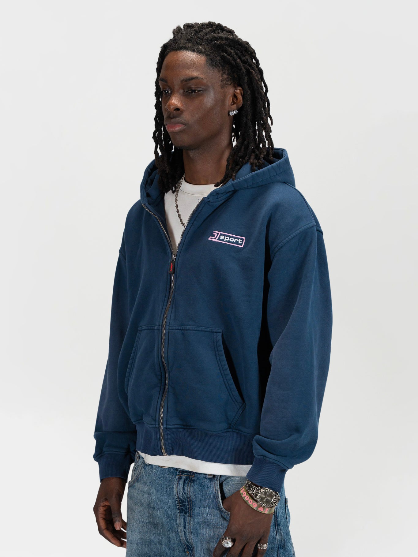 Front facing half body 45 degree shot of a male model wearing the J Sport zip by J3T. HE has on a white shirt underneath, light blue jeans and light brown boots to complete the fit.