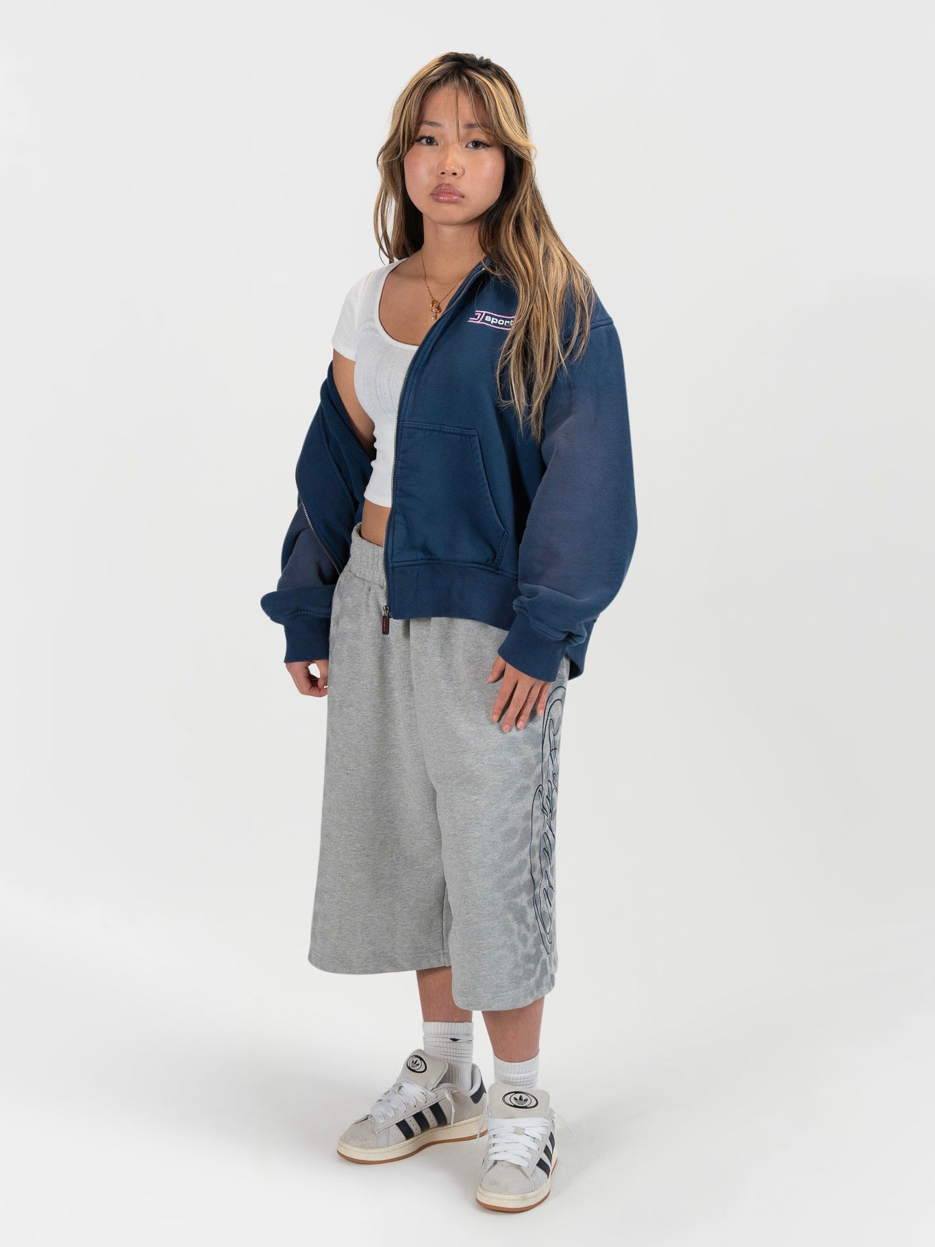 Front facing full body shot of a female model wearing the J Sport zip by J3T. she has on a white shirt underneath, the jafari shorts, and white sneakers to complete the fit
